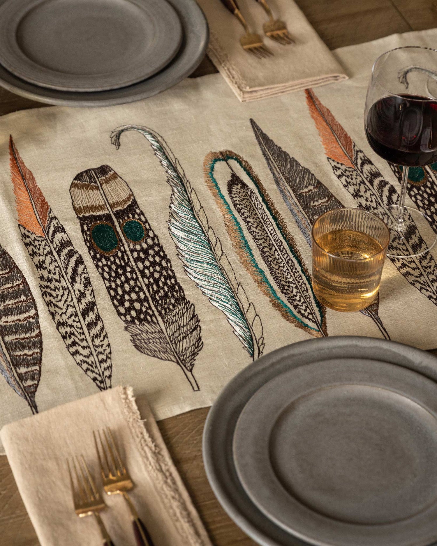 Overhead view of a dining table showing two table settings featuring Coral & Tusk embroidered linen Feathers Table Runner in the center, slate gray ceramic plates, glasses, and silverware on a wooden table.