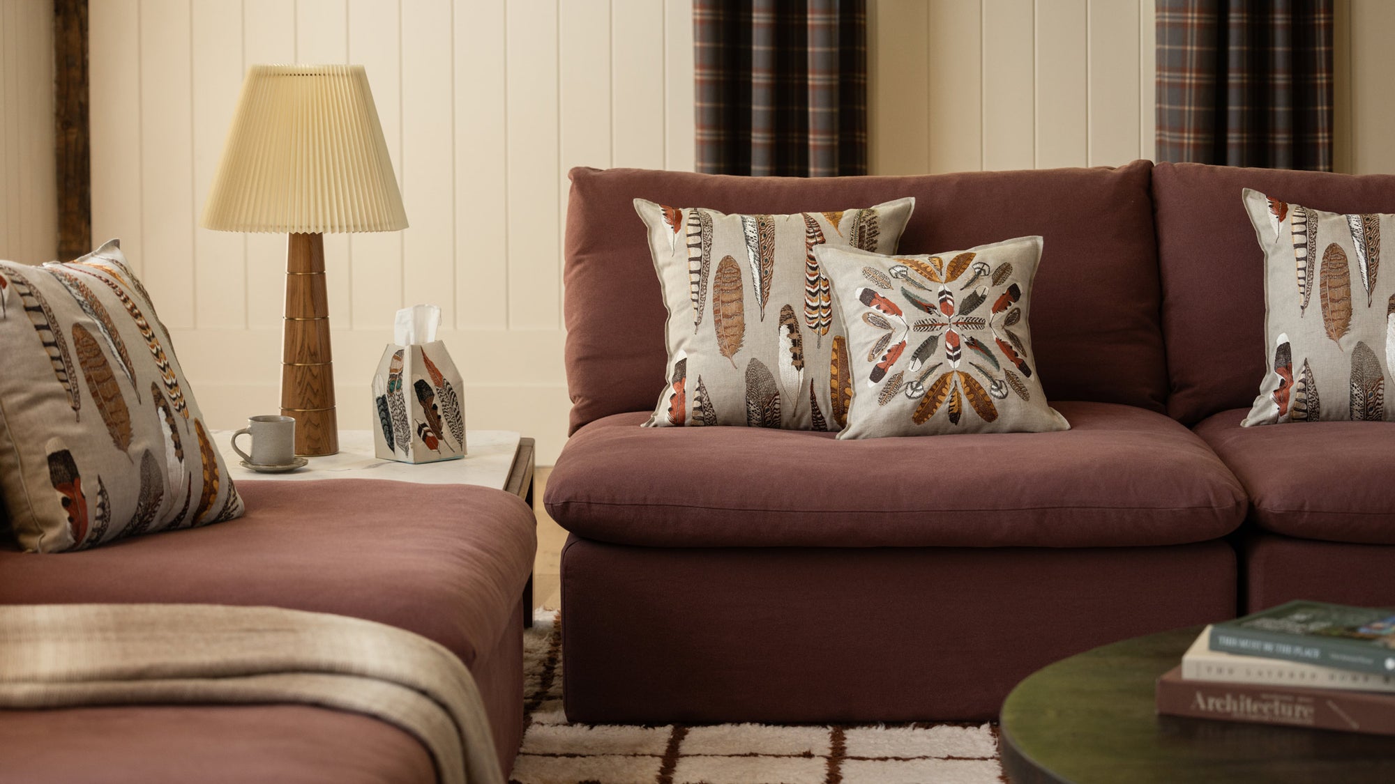 A styled living room scene featuring a maroon sectional sofa with four Coral & Tusk feather-themed decorative embroidered throw pillows, a side table with a lamp, a Coral & Tusk Feather Tissue Box Cover, and a coffee mug, and a round coffee table in the foreground with books.