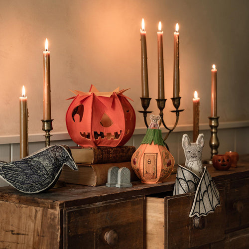 A styled Halloween-themed decorative display with Coral & Tusk objects, including embroidered pumpkins, a blackbird, a bat, and lit taper candles, on a wooden dresser against a beige wall.