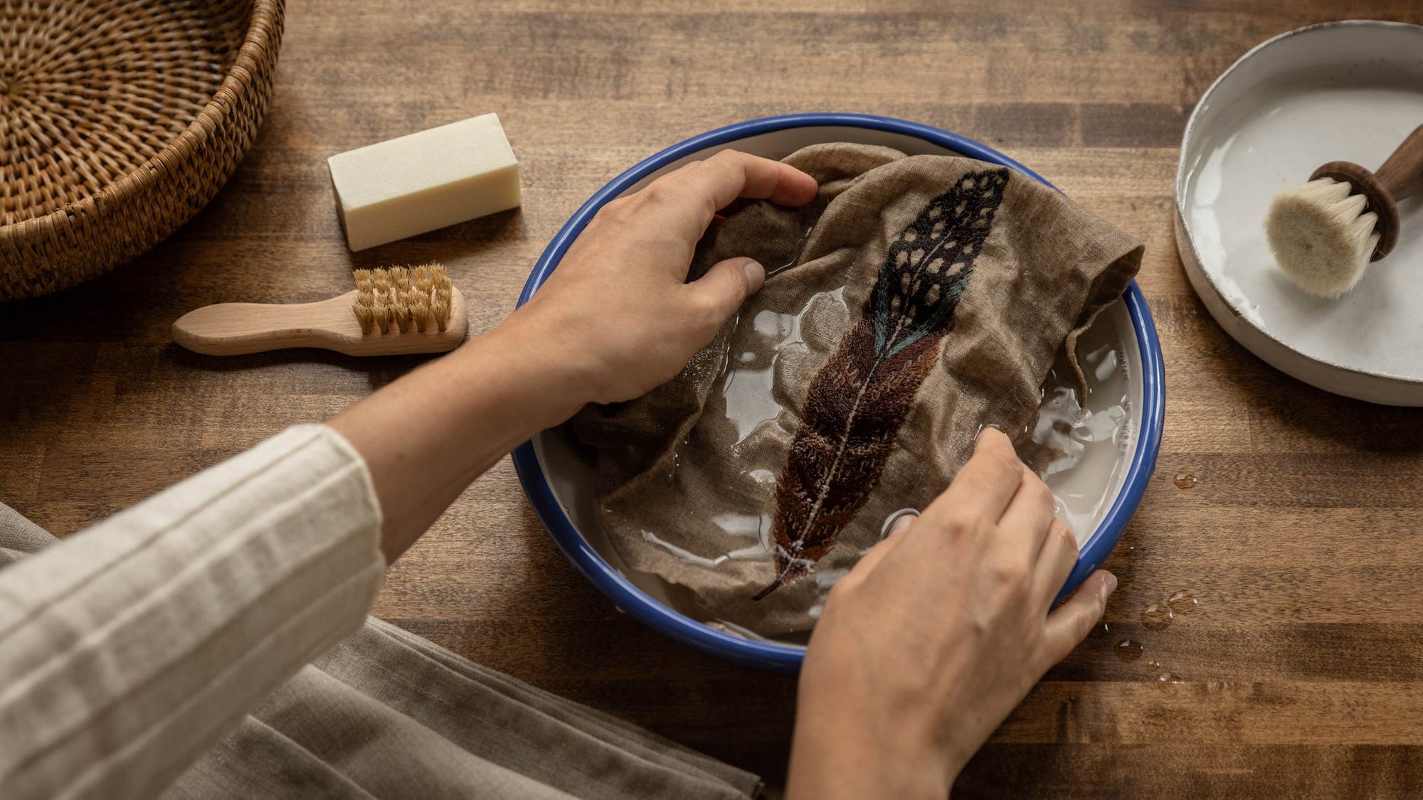 Overhead view of a person's hands washing a Coral & Tusk linen dinner napkin with a fowl feather embroidered on it in a blue-rimmed bowl on a wooden table, with a woven basket, a laundry spot-cleaning bar and brush in a dish beside the bowl, and a pile of linens beside it.