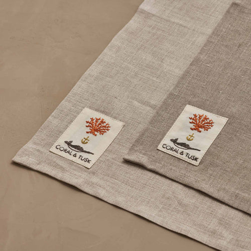 A close-up of Coral & Tusk dinner napkins, flat, showing the corners with the Coral & Tusk embroidered logo tag to compare natural variations in linen color. The top napkin is a slightly darker linen, though the embroidered tag and thread colors are the same.