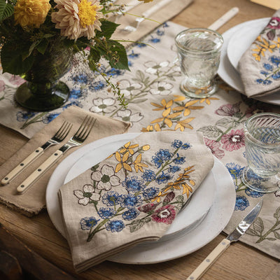 Table setting featuring Coral & Tusk Spring Stems embroidered Dinner Napkin and Table Runner, with glasses, white ceramic plates, silverware, and a bouquet of yellow flowers.