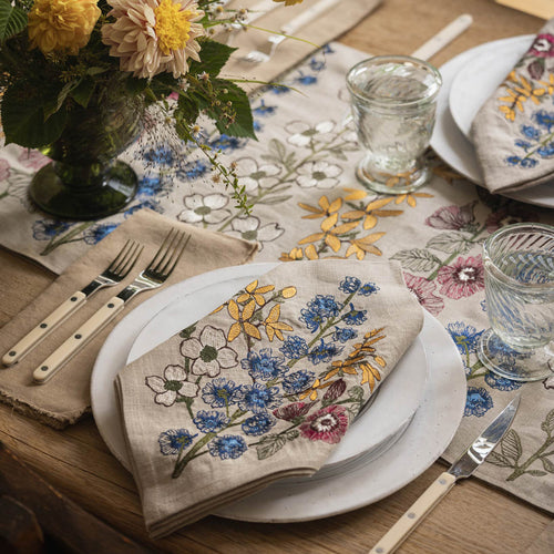 Table setting featuring Coral & Tusk Spring Stems embroidered Dinner Napkin and Table Runner, with glasses, white ceramic plates, silverware, and a bouquet of yellow flowers.