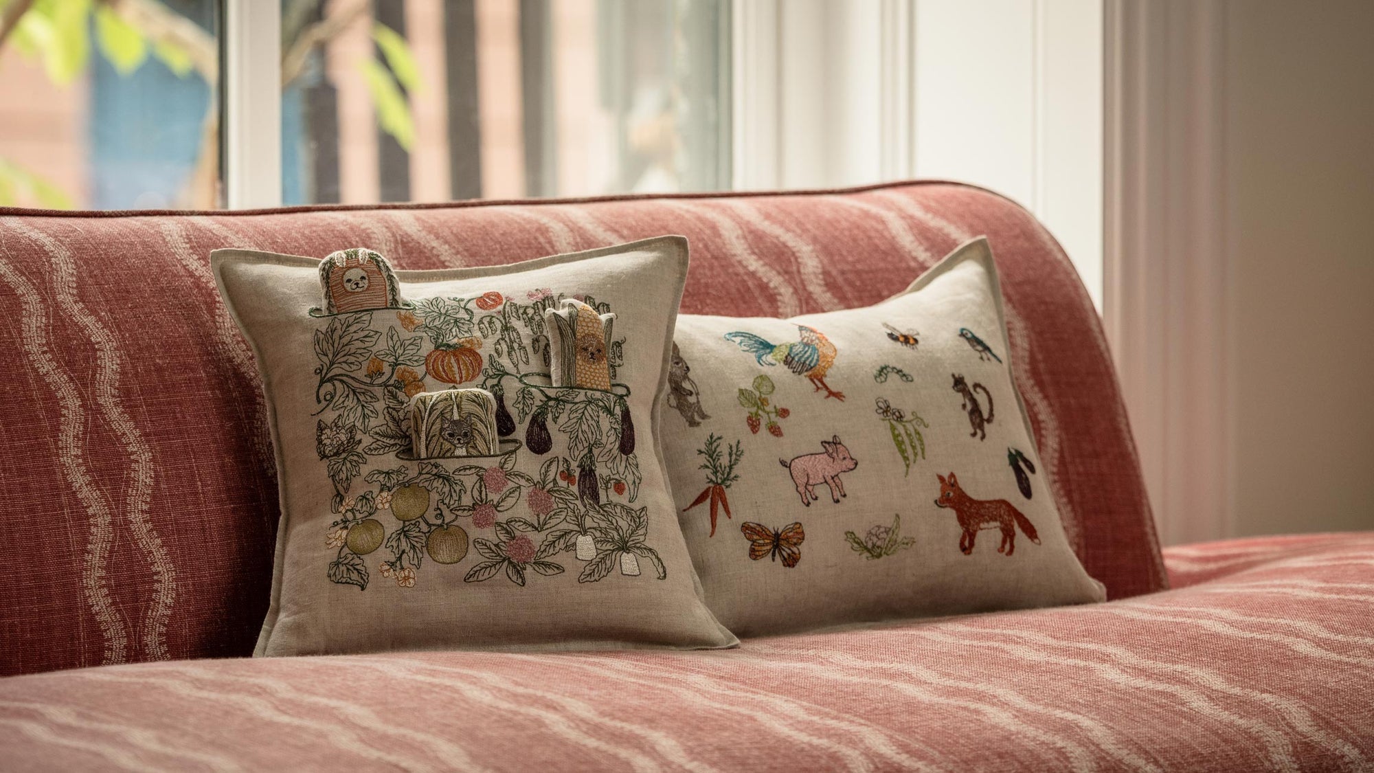 Coral & Tusk garden- and animal-themed embroidered decorative pillows on a red-striped sofa in front of a window with soft natural light.