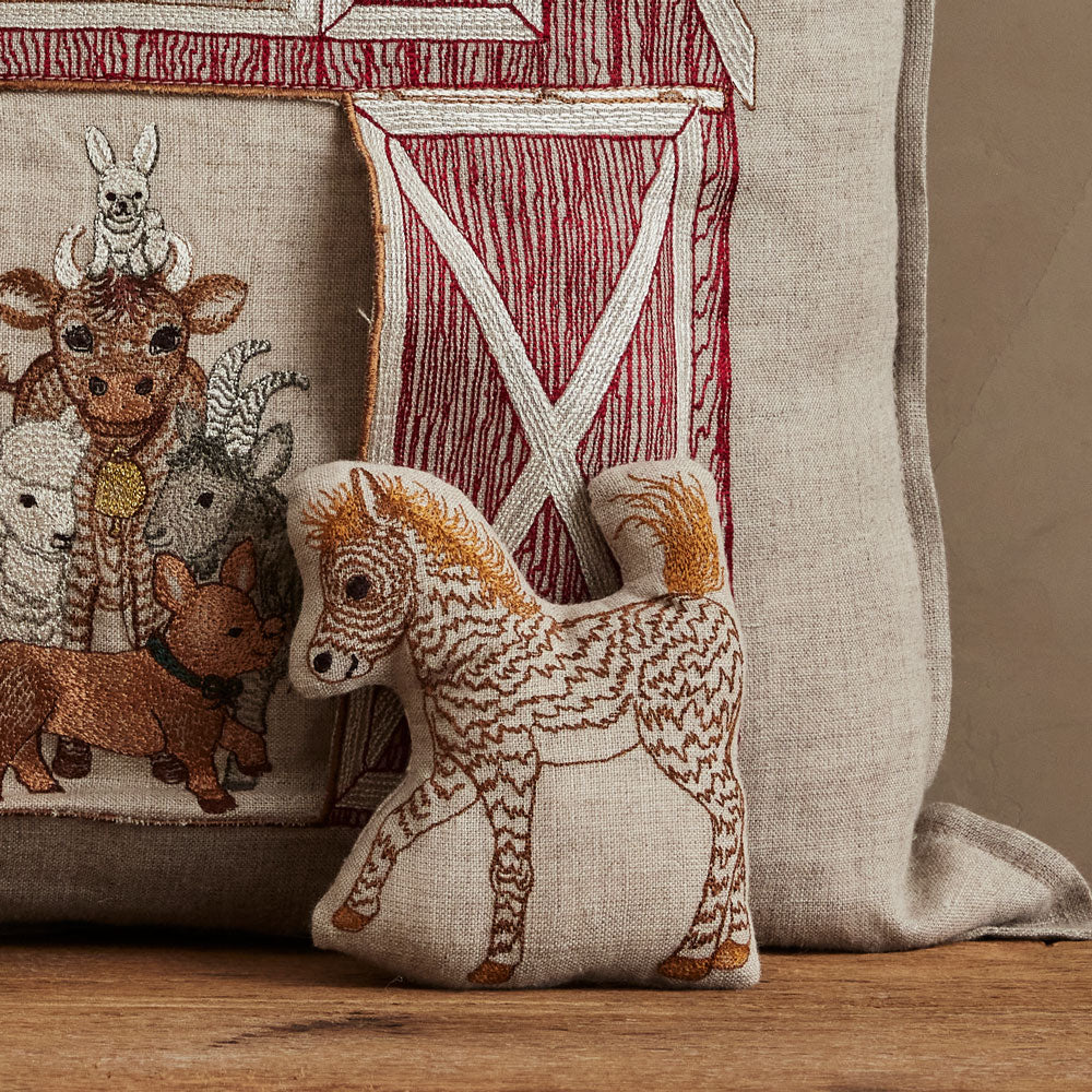 Close up of Coral & Tusk Red Barn Pocket Pillow with embroidered farm animal design and a horse pal on a wooden surface.