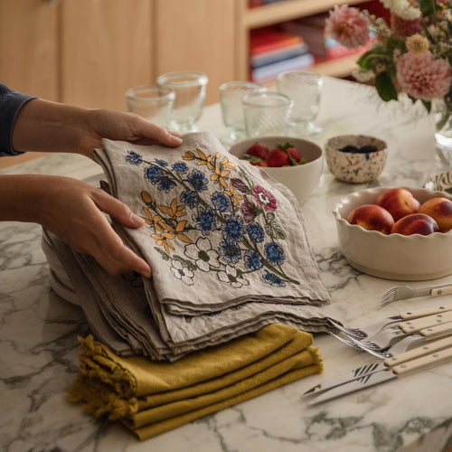 Hands placing a Coral & Tusk Spring Stems embroidered linen dinner napkin on a marble counter with stacked tableware, fruit, and flowers while setting a spring table for hosting.