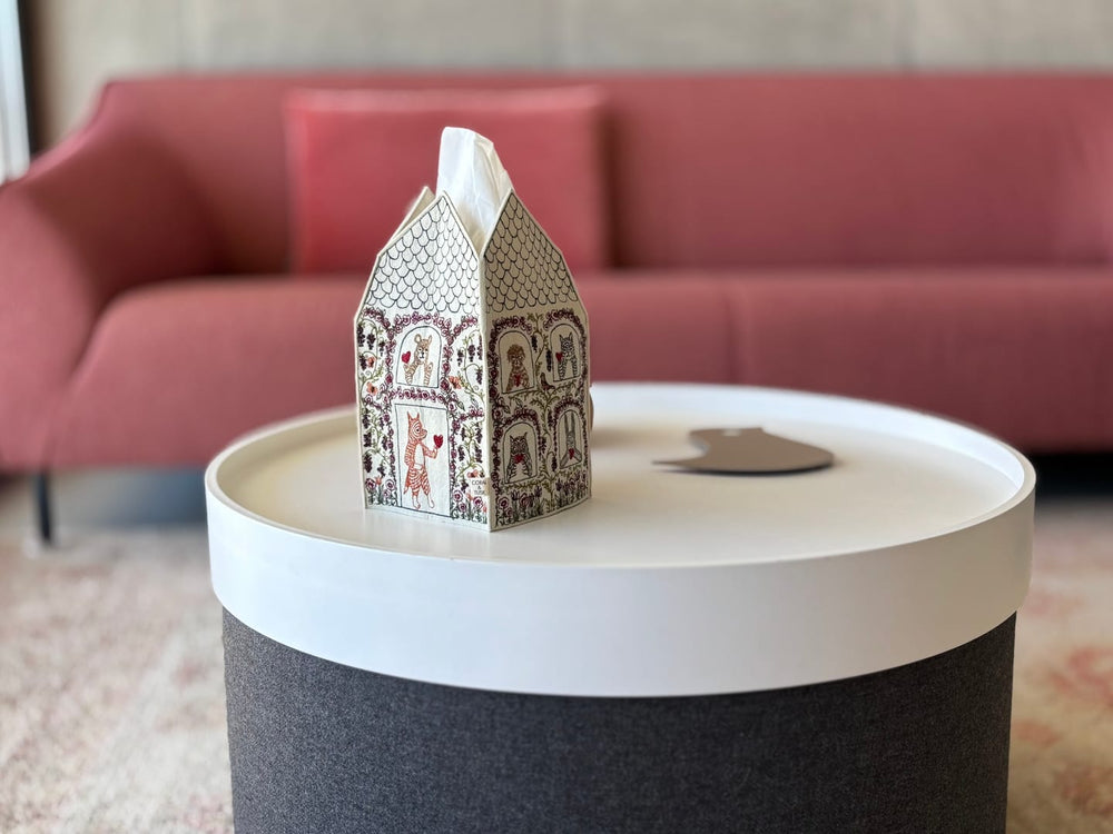 Decorative Coral & Tusk Tissue Box Cover featuring a house with woodland animals holding hearts in the door and window frames embroidered on all sides, sitting on a round table with a red sofa in the background.